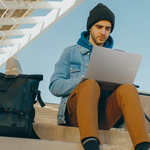 Young man sits on stairs outdoors with an ASUS Vivobook Pro 15 OLED laptop on his lap