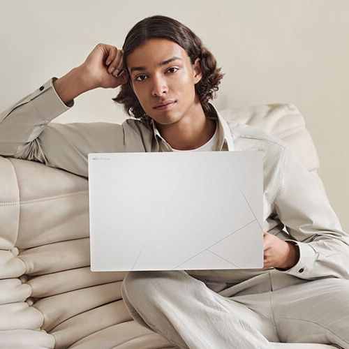 a young man sitting on a white sofa with a Zenbook S 16 laptop in Scandinavian White color on his lap
