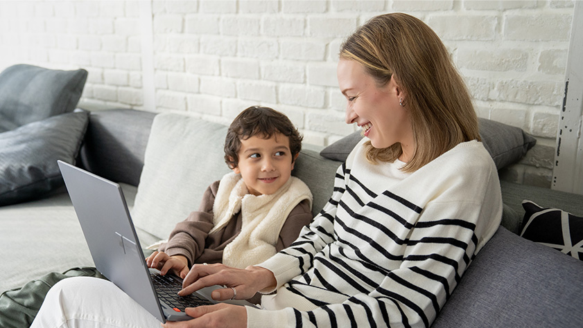 A boy and his mom uses one laptop together on the sofa. They press the keyboard at the same time and the boy smiles at his mom.
