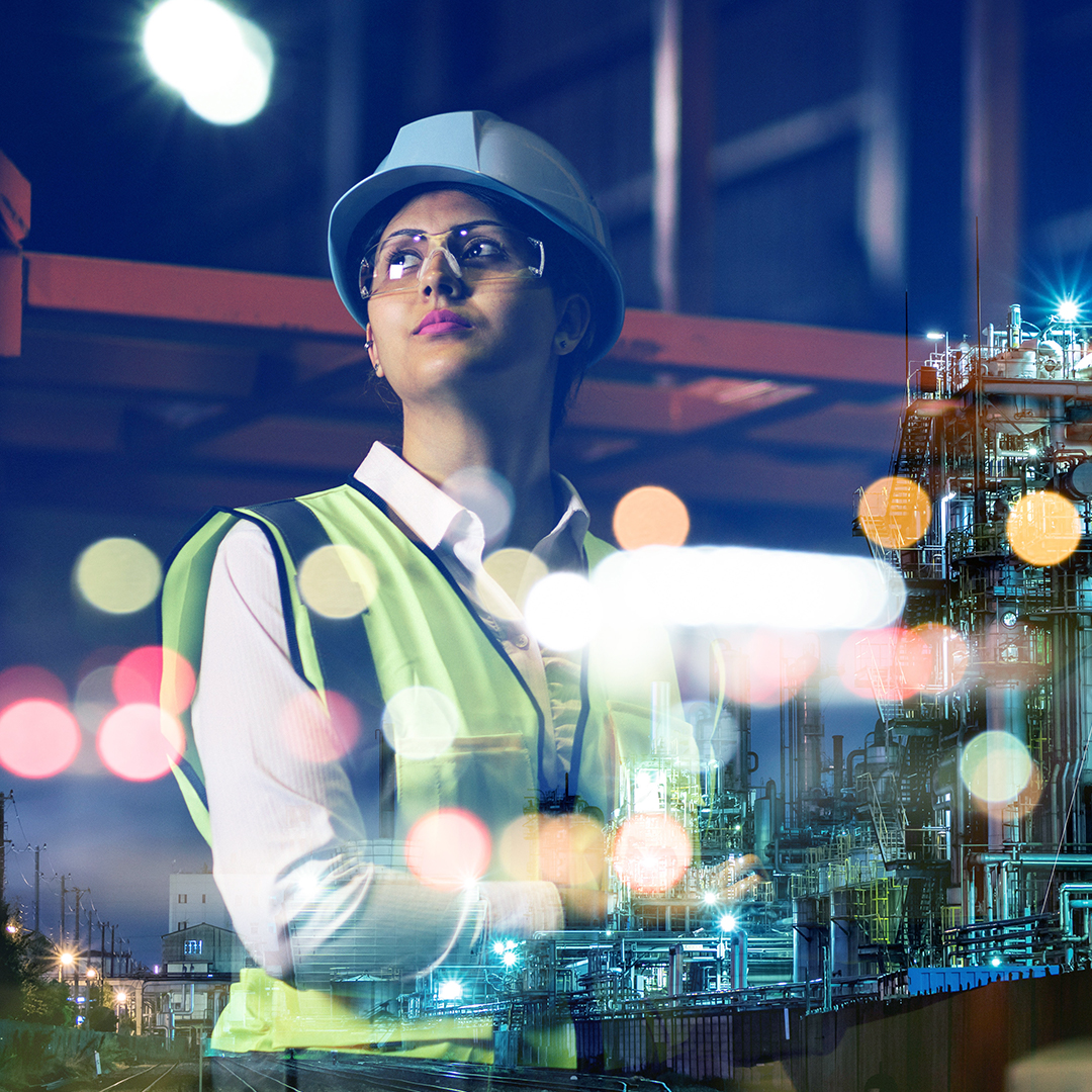 Female engineer wearing engineering hat and goggles looking ahead with large chemical factory in background