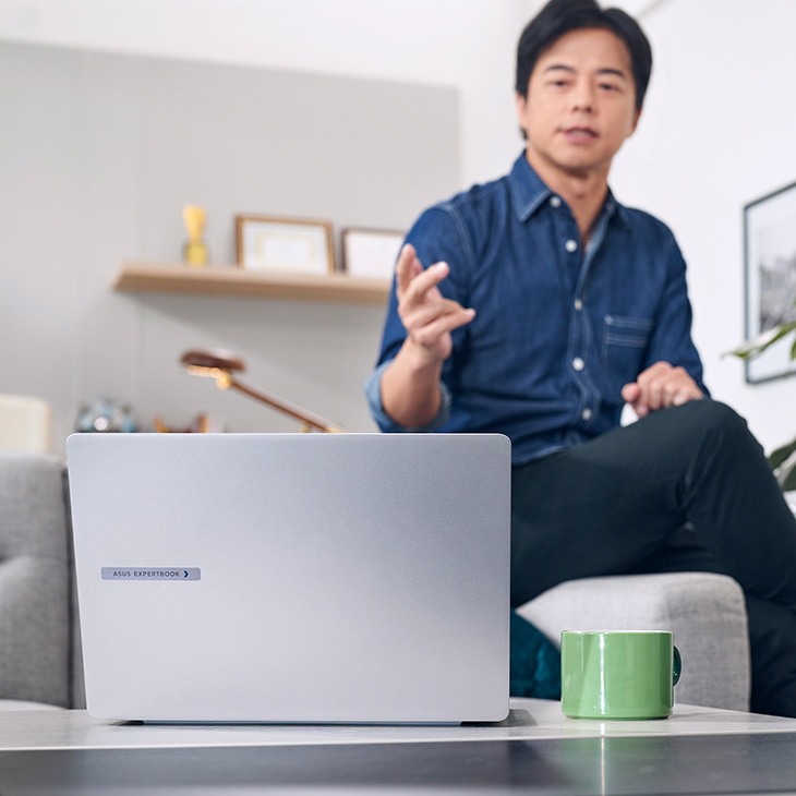 A business manager is sitting on a couch and having an online meeting in a modern home office. In front of him on a table, there is a silver ExpertBook AI laptop.