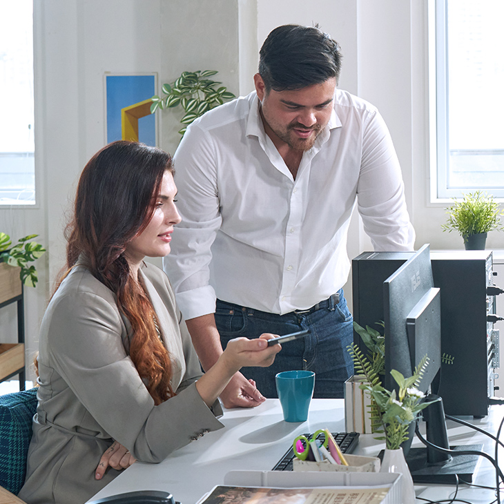 Two employees in a modern office setting. The woman is seated at a desk, holding a pen and pointing at the ASUS ExpertCenter desktop monitor. The other man is standing beside her, looking at the monitor.