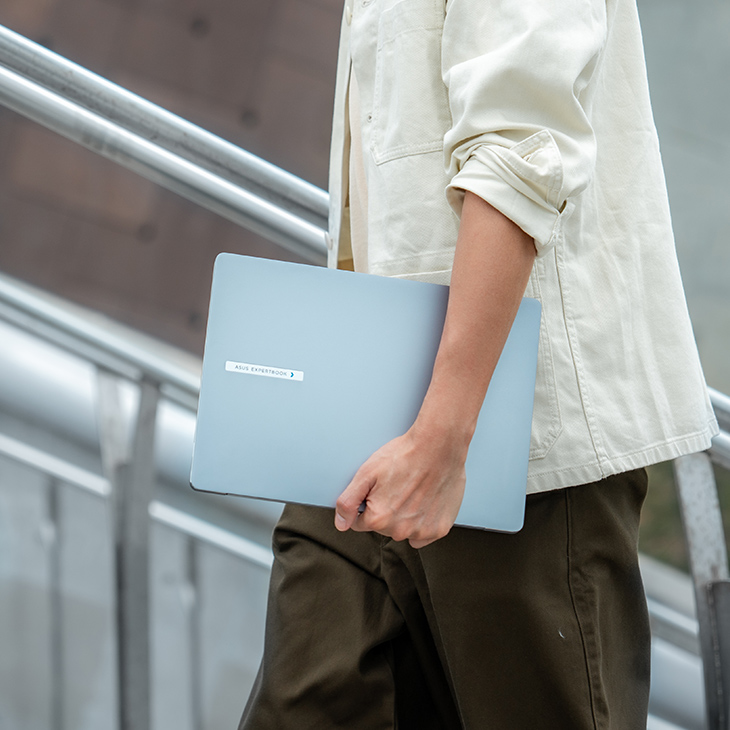 A young man is walking up a staircase, holding a closed ASUS ExpertBook P5 AI laptop in his right hand.