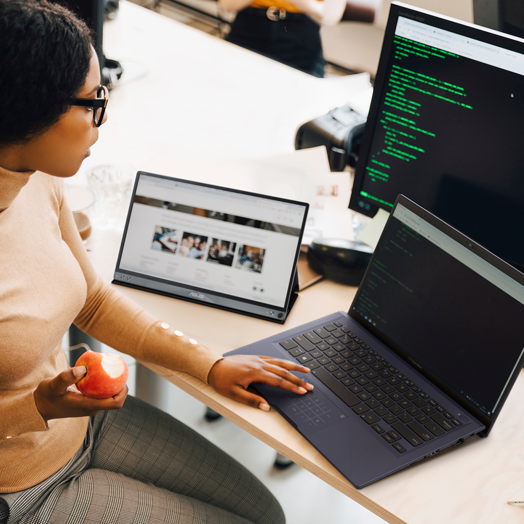 Una mujer del personal de TI está trabajando con una computadora portátil ExpertBook que se conecta con un monitor portátil ZenScreen y un monitor normal en el escritorio.