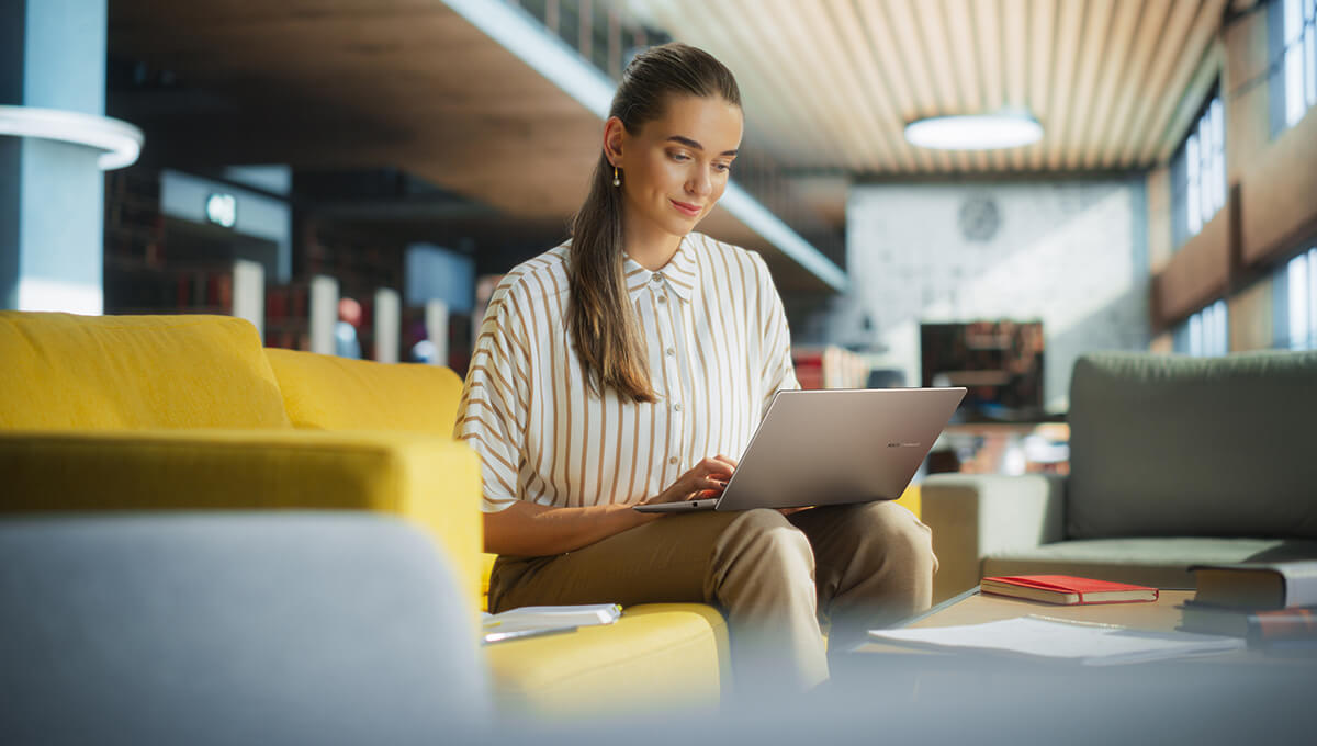 A woman is using a silver ASUS Vivobook on a yellow sofa in a lounge setting.