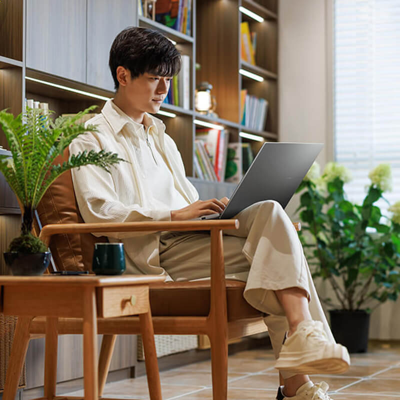 A young man is sitting in a chair using ASUS Vivobook S16 with several plants in the background.