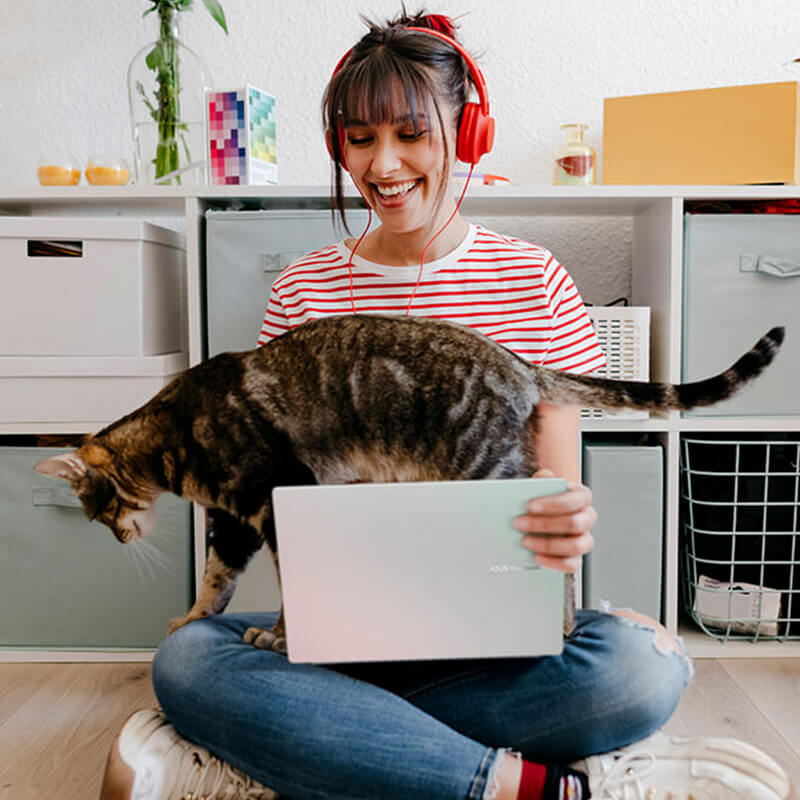 A young woman wearing red headphones sits on the floor holding an ASUS Vivobook 14 laptop on her lap while a cat walks past its keyboard