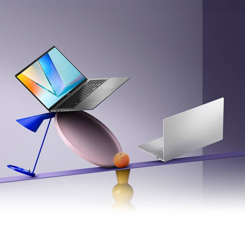 A Matte Gray ASUS Vivobook S16 and a Silver ASUS Vivobook S16 are balanced on a colorful, artistic seesaw with geometric shapes set against a soft purple gradient background.