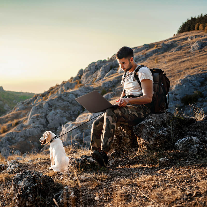 A man is sitting on a rock by a hiking trail with a ProArt laptop on his lap. A white Labrador is sitting in front of him looking out at the mountain scenery.