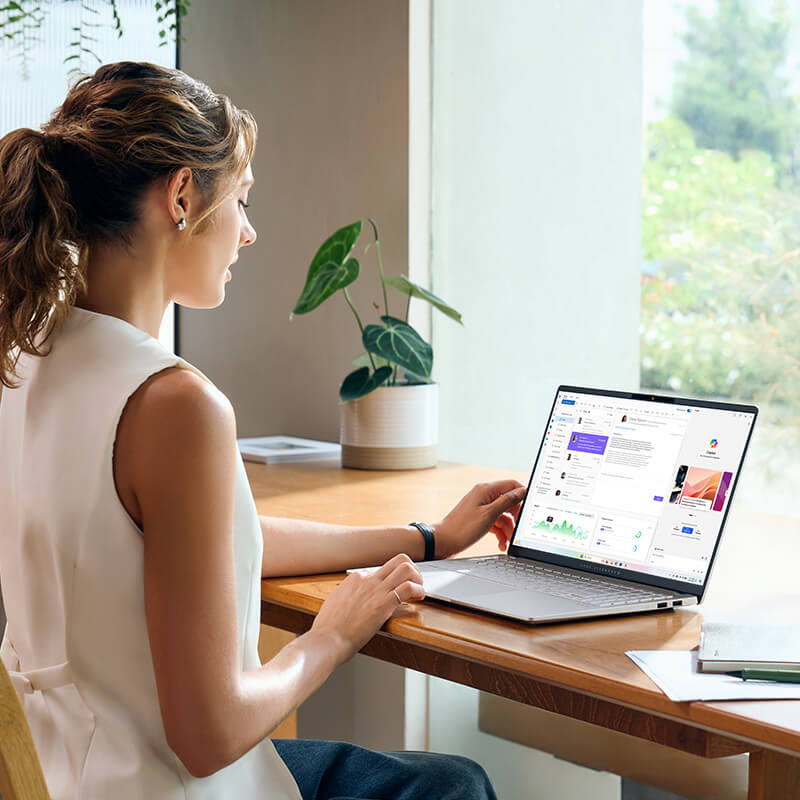 een jonge vrouw zit aan een bureau bij een raam met haar ASUS Vivobook S 15 laptop terwijl ze de AI-tool Copilot gebruikt