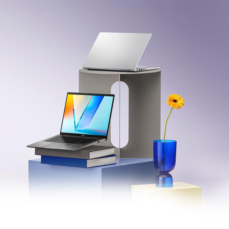 Two open laptops sit on a geometric stand. The black laptop is hovering above two books while a blue vase with a yellow flower sits on the right.