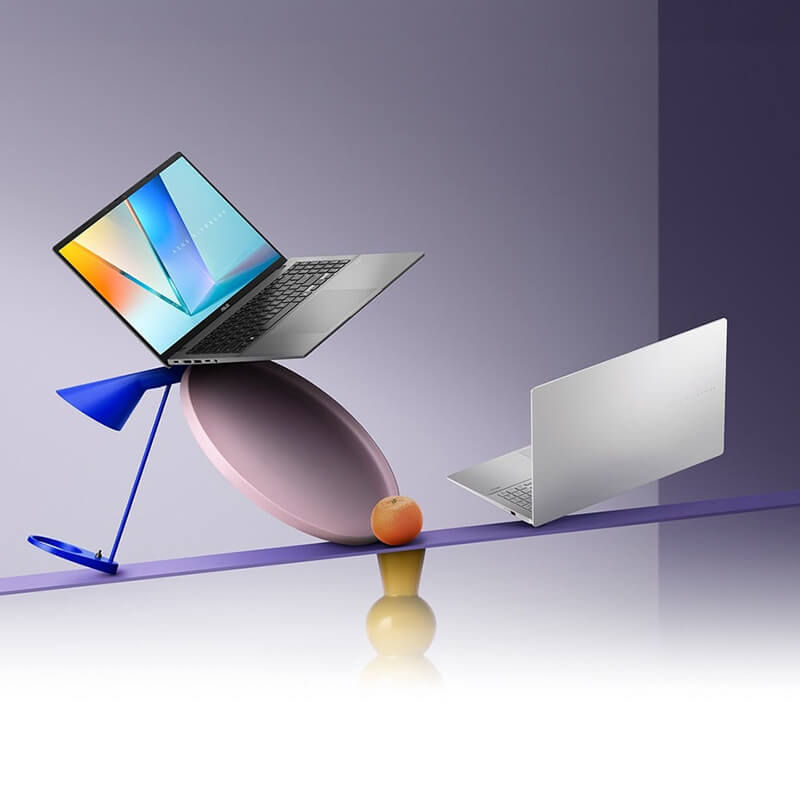 A Matte Gray ASUS Vivobook S16 is open while a Cool Silver ASUS Vivobook S16 is closed. Both laptops are balanced on a colorful, artistic seesaw with geometric shapes set against a soft purple gradient background.