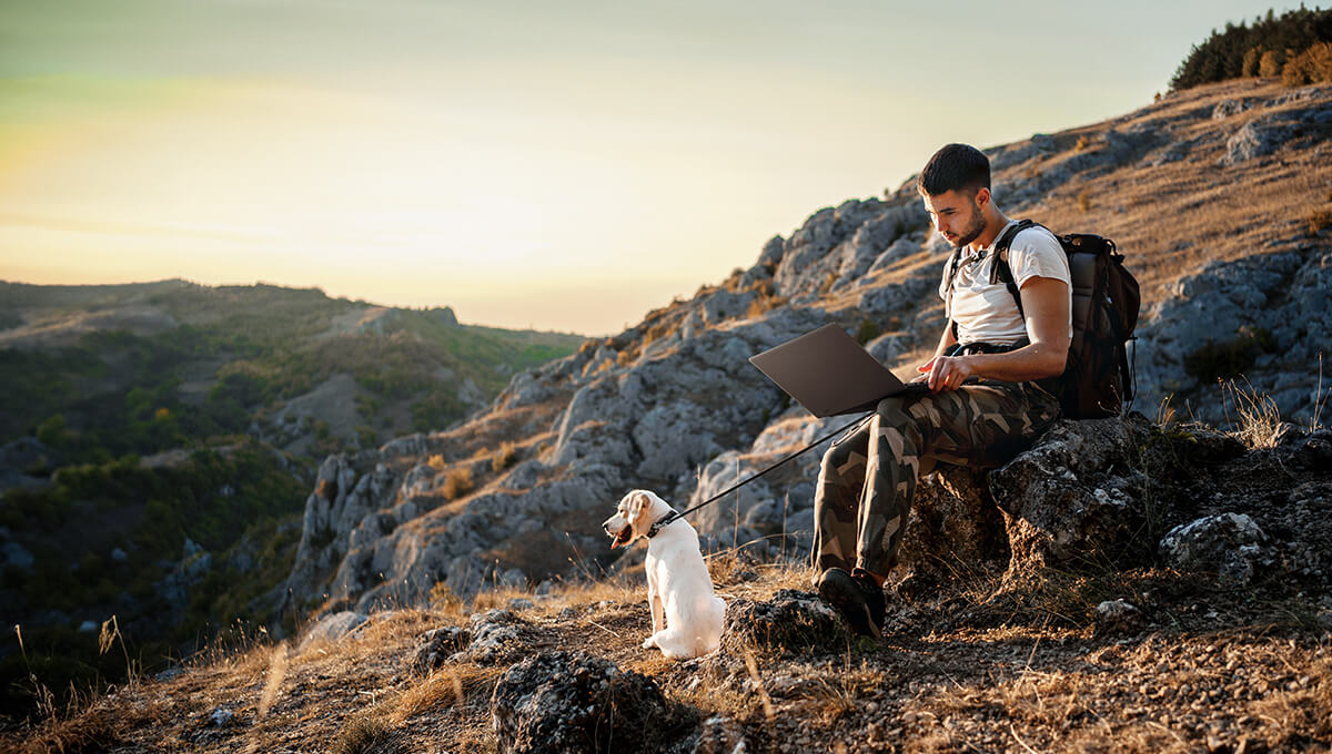 A man is sitting on a rock by a hiking trail with a ProArt laptop on his lap. A white Labrador is sitting in front of him looking out at the mountain scenery.