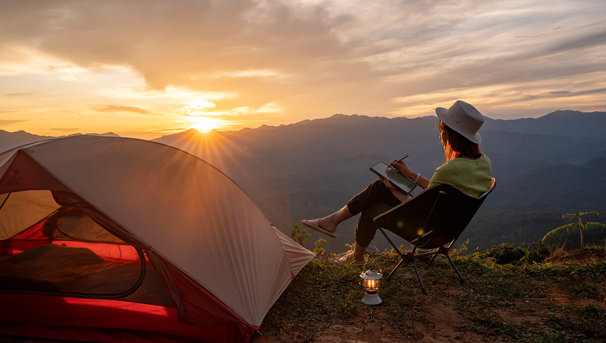 A woman is using the ASUS Pen 2.0 stylus on the ProArt PZ13 laptop beside her tent. She is sitting by her red tent on the mountains overlooking the setting sun.