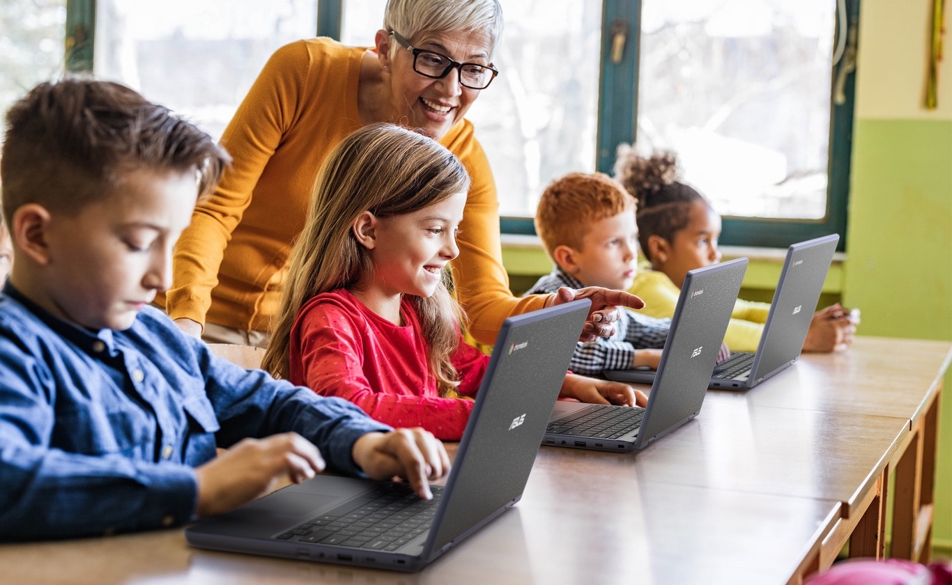 Driemaal schuin achteraanzicht van de ASUS Chromebook CR11 Flip in laptopmodus op een bureau. Drie kinderen gebruiken ze voor de les en een vrouwelijke leerkracht staat achterin de klas om hen te ondersteunen. Een ander meisje op de laatste rij kijkt naar voren.