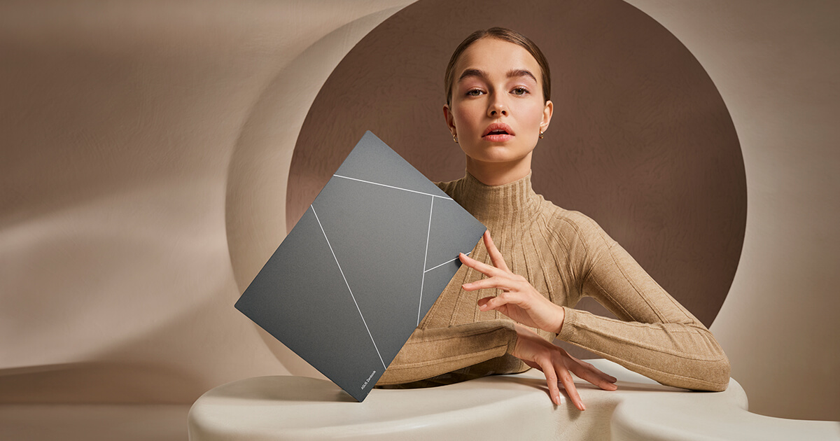 A woman is holding Zenbook S 13 OLED on its corner with her fingertips. The Zenbook monogram on the basalt grey lid is displayed as the subject of the image.
