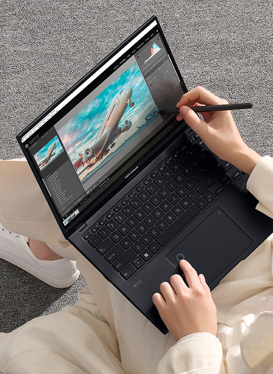 A woman holds a stylus to work on Zenbook Pro 14 OLED’s screen with her right hand. There is airplane photo shown on the in-screen. And there is a camera placed next to the women on the bench she is sitting on. 