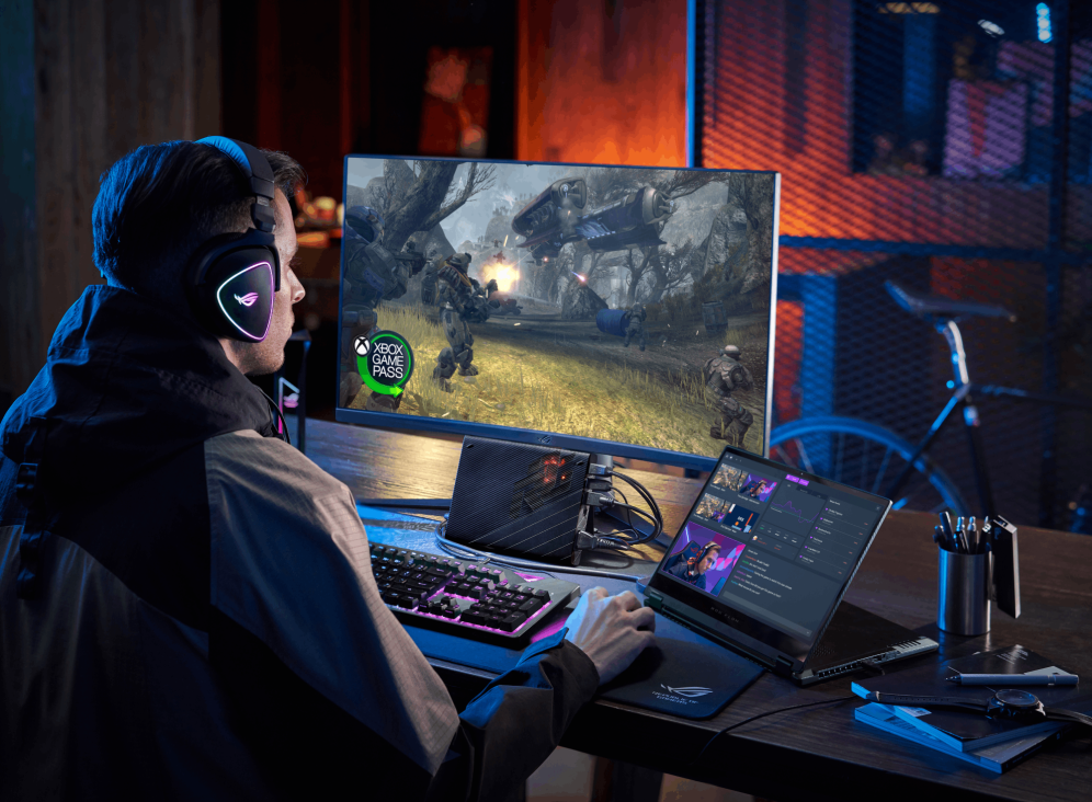 Gamer at a desk with a bike in the background, and a Flow X13 in stand mode connected to a headset, mouse, keyboard, XG Mobile, and a monitor.