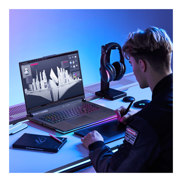 A person sitting at a desk with their back to the camera, working on a drawing pad, with a Strix SCAR 18 open in front of them, and with a mouse, headphones, and notepad on the desk