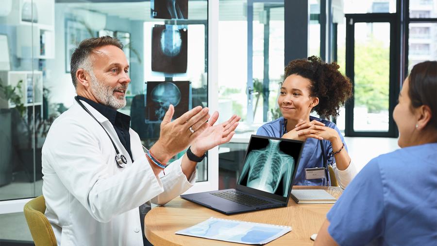 Valkoiseen lääkärintakkiin pukeutunut mies ja kaksi vaaleansiniseen sairaanhoitajan asuun pukeutunutta naista istuvat puisen pöydän ääressä. Pöydällä on auki ExpertBook -kannettava, jonka näytöllä näkyy röntgenkuva ihmiskehosta. 