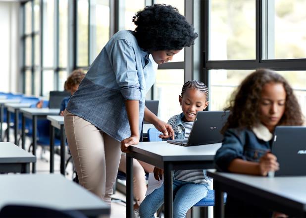 Anak-anak menggunakan laptop ASUS BR1100 mereka di dalam kelas dan seorang guru sedang mengajarkan seorang anak perempuan, mereka melihat laptop bersama-sama.