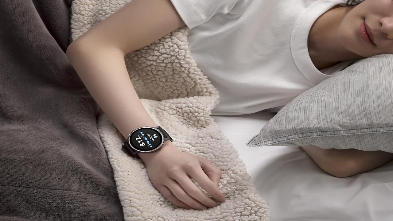 A person sleeps on a bed wearing an ASUS VivoWatch 6 smartwatch displaying the time and sleep data. The scene conveys comfort with soft bedding and neutral colors.