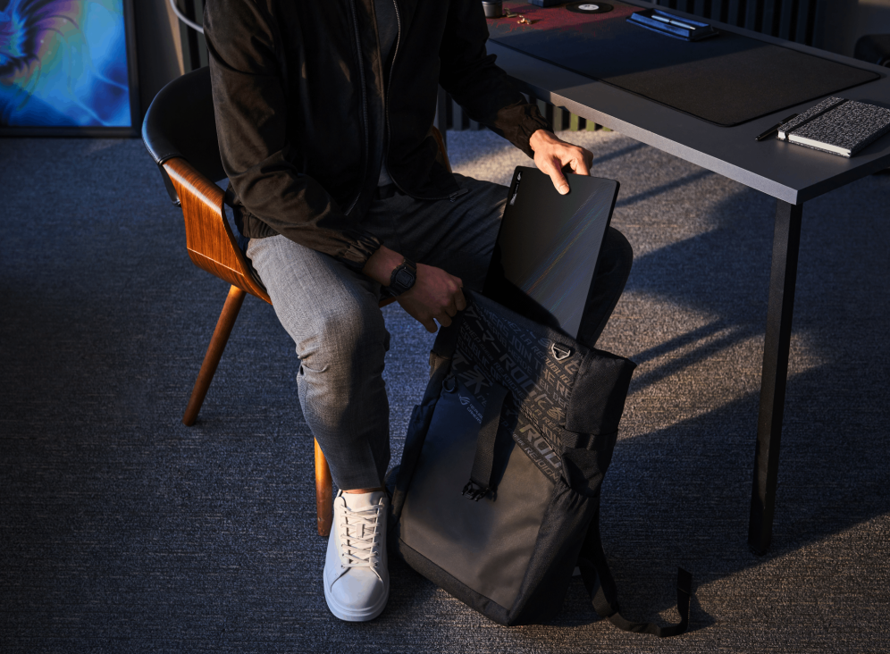 Person seated at a table, slipping a black Zephyrus M16 into an ROG backpack.