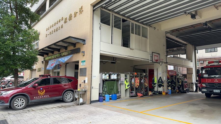 Fire station with open garage, housing a fire truck and firefighter gear. A red SUV with a fire department emblem is parked outside. Urban setting.