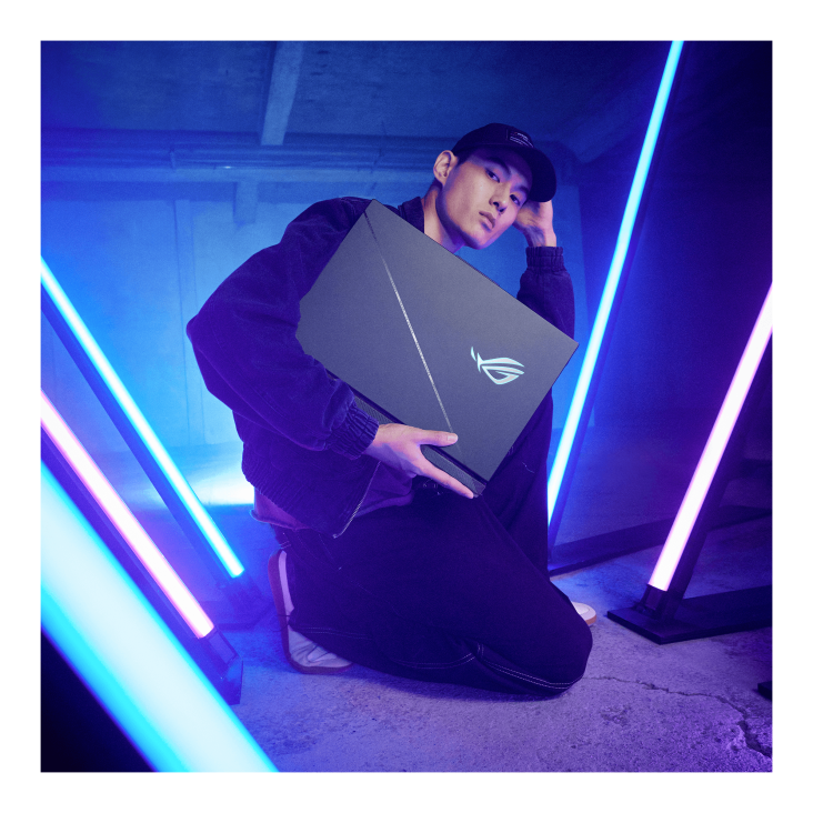 A person sitting in the middle of some neon lights, looking at the camera, and proudly displaying a Strix SCAR gaming laptop