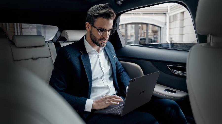 A man working on ASUS ExpertBook B9 OLED in the back seat of a luxury car, wearing a suit and the background is a city street.