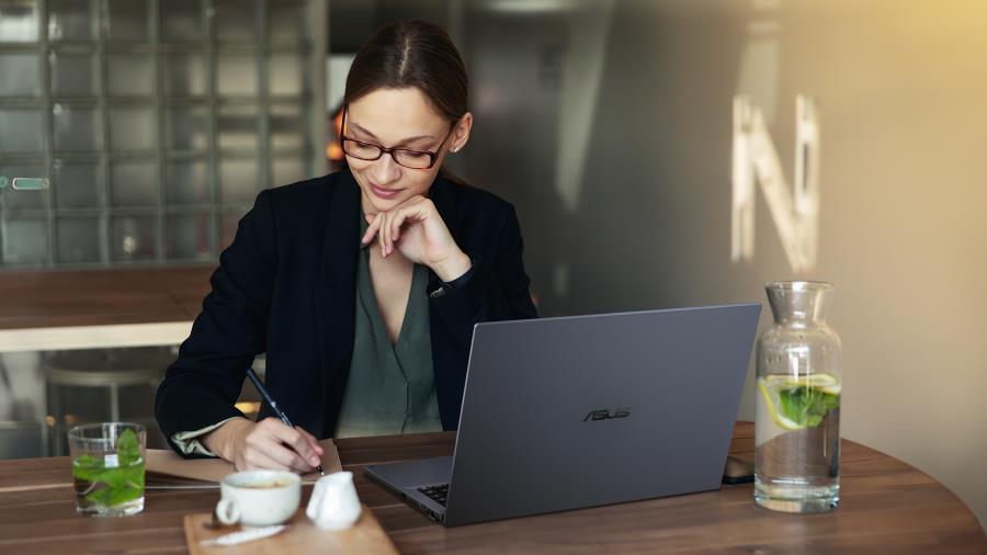 A woman working on an ASUS ExpertBook B2  in a cafe. She is wearing a black blazer and have a glass of water with lime and a cup of coffee on the table.