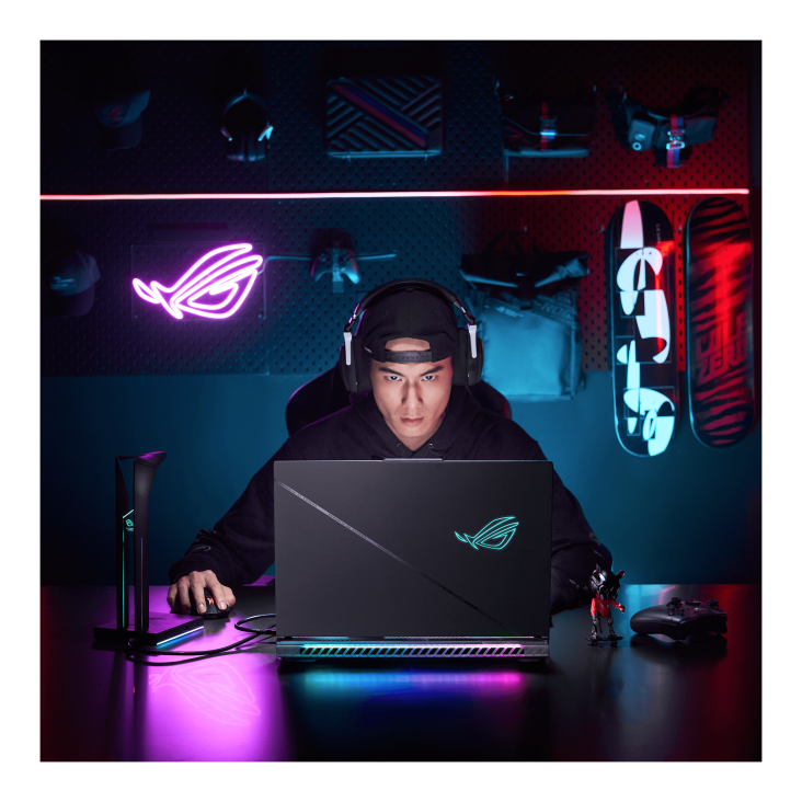 A dark room with purple lighting, a backpack, skateboards, and an ROG neon sign on the wall, with a gamer facing the camera and wearing a headset while using the ROG Strix SCAR laptop