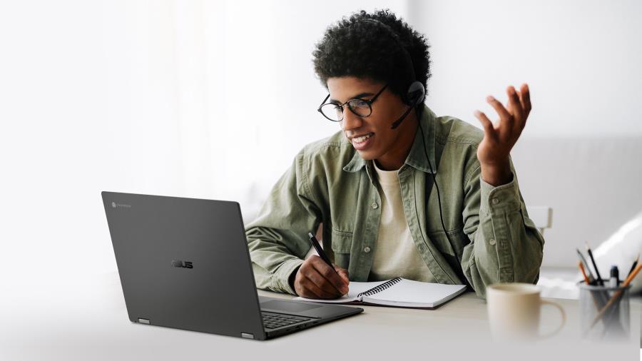 An african man with glasses and earphones and a microphone is taking notes with an ASUS Chromebook CX5 in front of him.