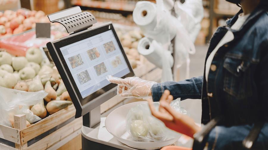 A masked customer using a self-checkout machine to purchase fruit, wearing gloves, and interacting with a touchable panel.