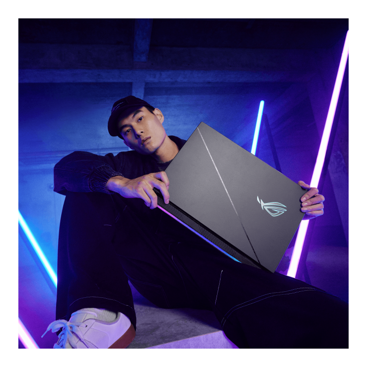 A person sitting on a concrete block, looking at the camera, and proudly displaying a Strix SCAR gaming laptop
