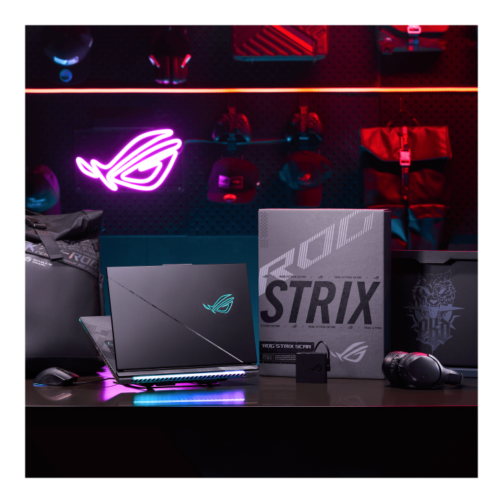 A dark room with purple lighting, with a desk featuring an ROG backpack, mouse, Strix SCAR gaming laptop, and the packaging the laptop comes in
