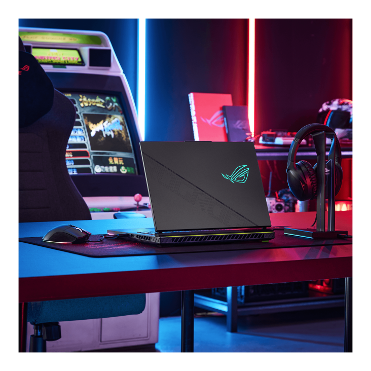 A game room, with an arcade machine in the background, with a desk featuring a Strix G laptop, a mouse, and a headset, as well as an ROG gaming chair in the foreground