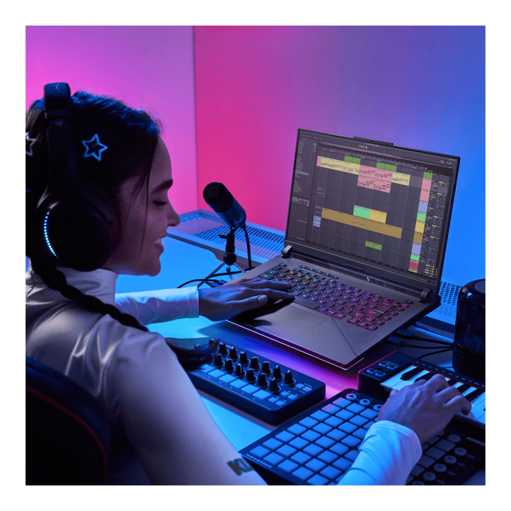 A view over the shoulder of a person working on a music track on an ROG Strix G laptop, wearing headphones, with a microphone, keyboard, and digital music controllers also visable