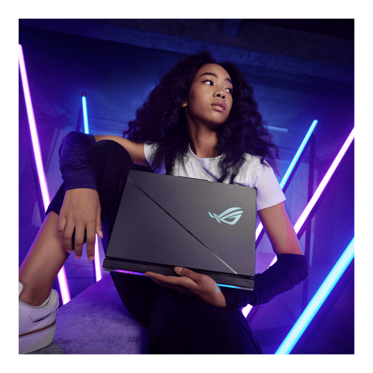A person sitting on a concrete block with neon lights in the background, looking away from the camera, and proudly displaying a Strix SCAR gaming laptop
