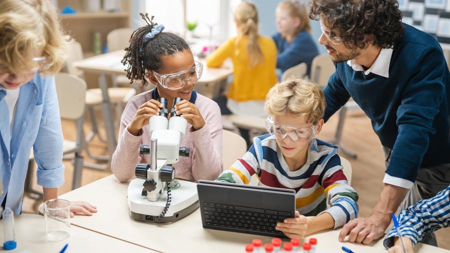 A teacher with kids in a classroom, one boy wearing eye protector is using ASUS laptop in tablet mode and one girl wearing eye protector is using microscope. Others are chatting.
