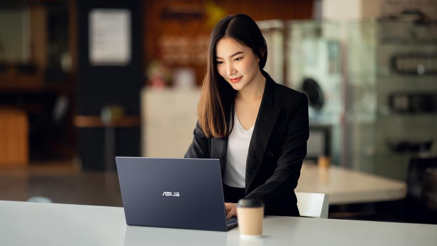 A woman sitting at a desk with an ASUS ExpertBook B1 laptop and a white coffee cup. The desk is wooden, and the background features a modern office space with a blackboard and a plant.