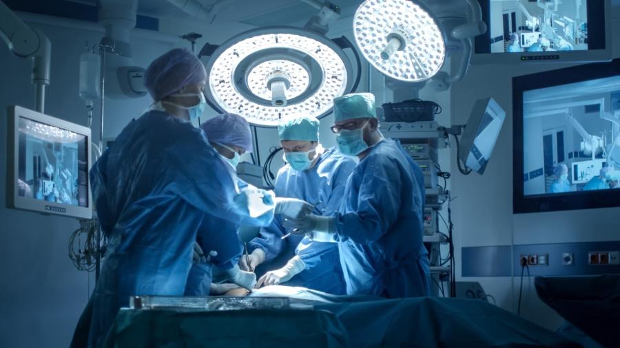 A doctor in the operating room, accompanied by three medical staff members performing simultaneous procedures, surrounded by three screens.