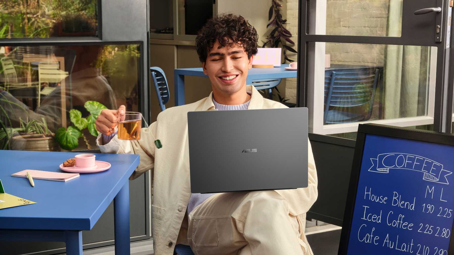 A student uses an ASUS Vivobook S16 laptop while sitting at a table in a modern, brightly lit café.