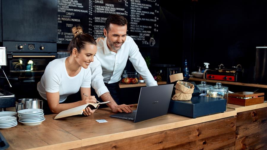 A male cafe owner using the ASUS ExpertBook L1 in a cafe behind the counter, with the female owner on his right writing on a notebook.
