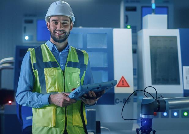 A man in a factory holds a tablet and stands in front of the machine