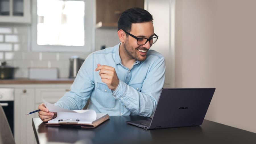 A man with a beard and glasses is holding a pen and a pieice of paper in his right hand smiling to the screen with an ASUS ExpertBook B3 Flip in front of him. 